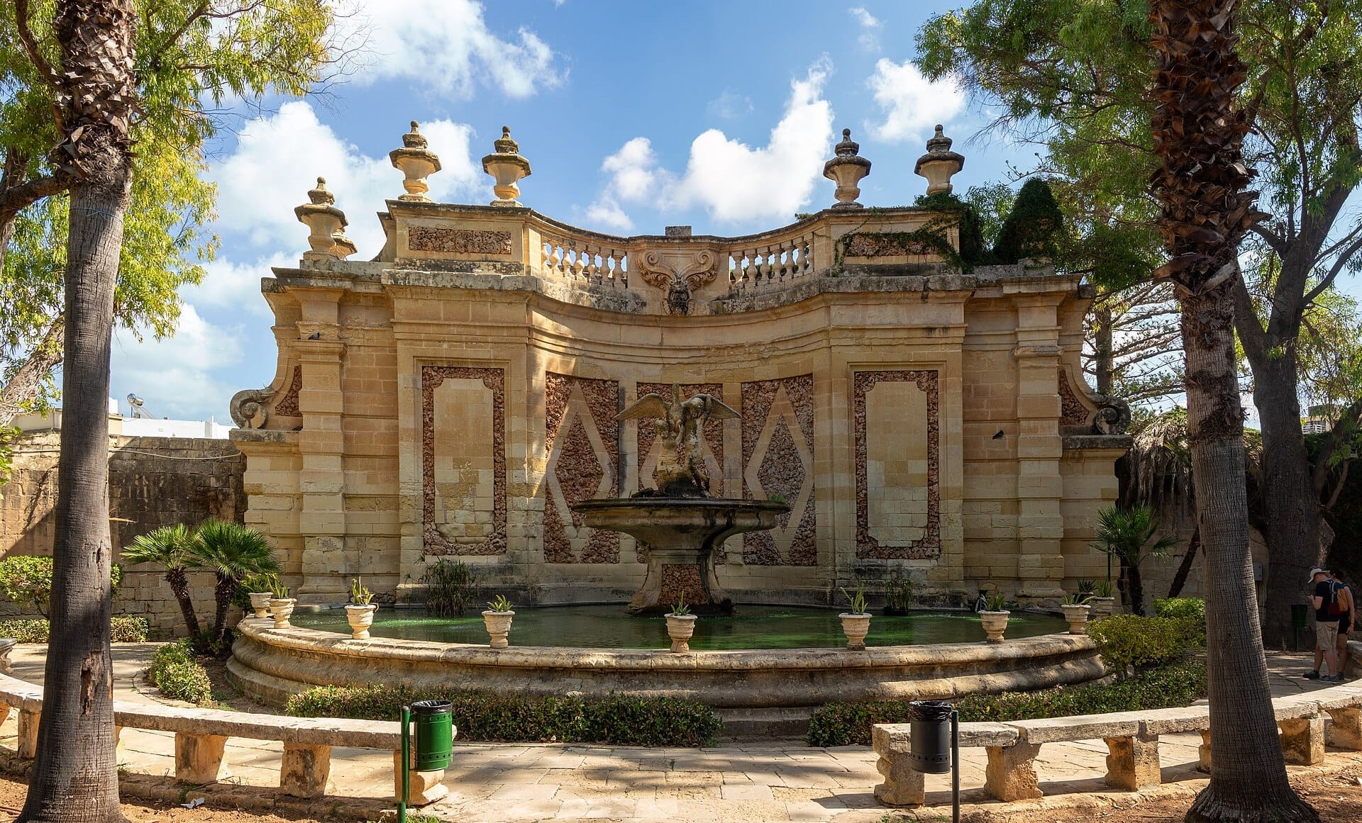 Fontana centrale con una statua alata, circondata da un'architettura storica in arenaria e una rigogliosa vegetazione nei San Anton Gardens alla luce del giorno.