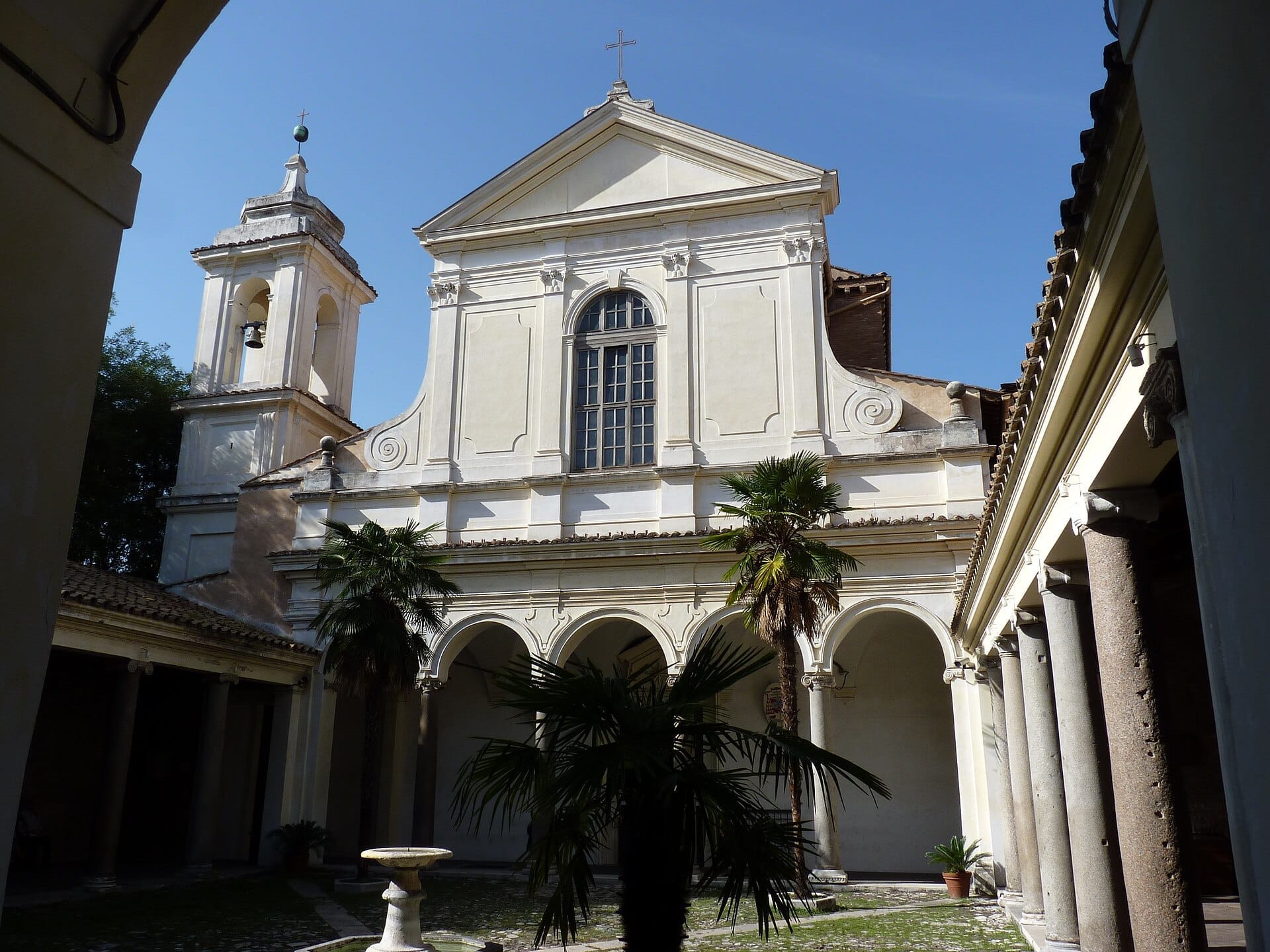 San Clemente Basilica's cream-colored facade, arched cloister, and bell tower seen on a sunny day in Rome.