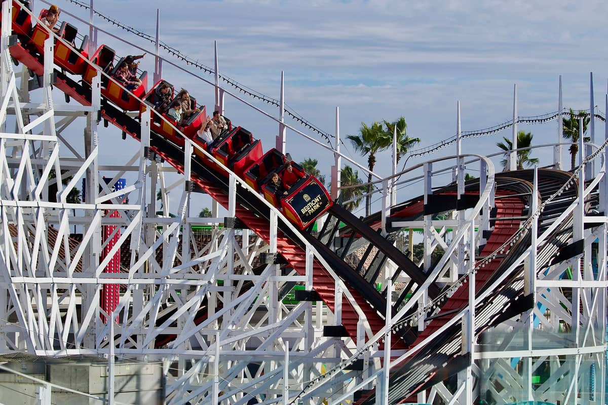 ภาพระยะใกล้ของรถไฟเหาะ Giant Dipper ที่ Belmont Park ใน San Diego มีผู้โดยสารในรถสีแดงและรางไม้สีขาว พร้อมต้นปาล์มในพื้นหลัง
