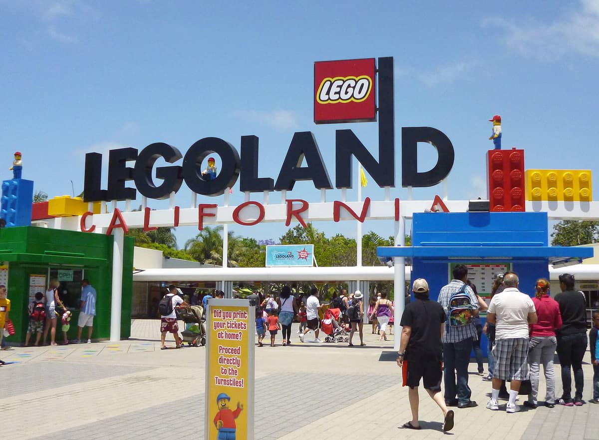 ทางเข้าหลักของ LEGOLAND California ในวันที่แดดจัด พร้อมกลุ่มครอบครัวและเด็กๆ ที่กำลังเข้าสวนสนุก โครงสร้างสีสันสดใสในธีม LEGO และป้ายด้านบน