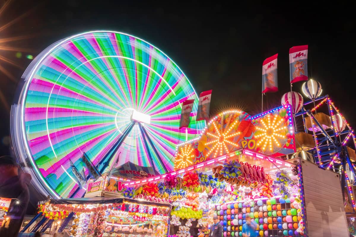 Buntes Riesenrad und lebhafte Spielbuden auf einem großen Jahrmarkt bei Nacht beleuchtet.
