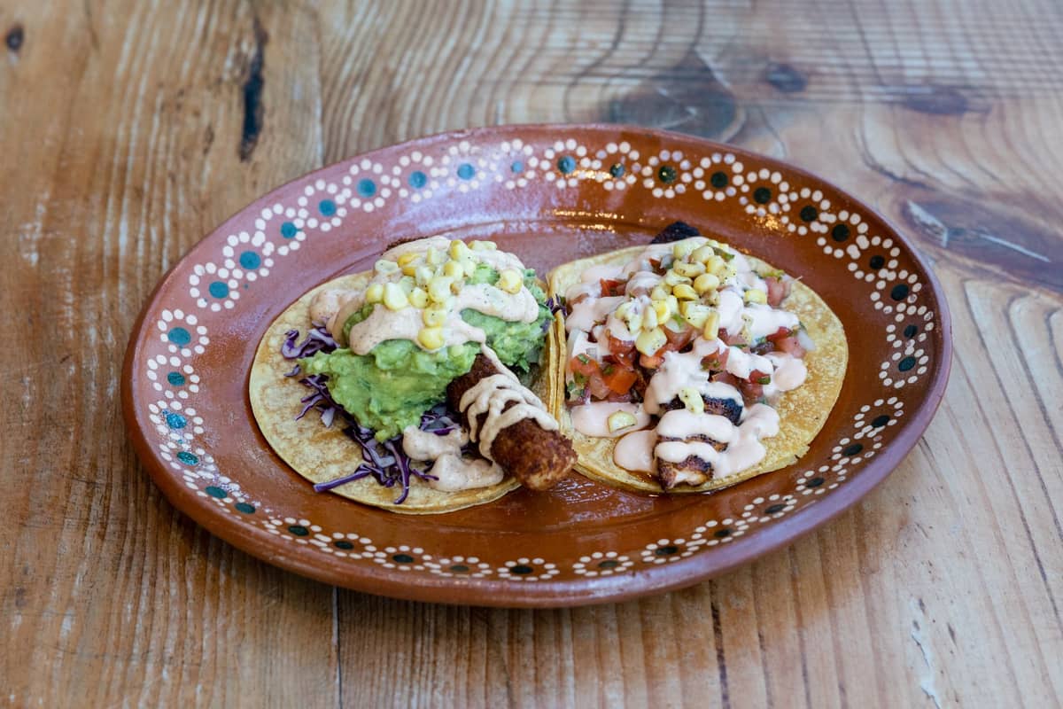 Zwei Fish Tacos mit frischen Toppings wie Guacamole, cremiger Sauce und Mais auf einem rustikalen Holztisch, Nahaufnahme.