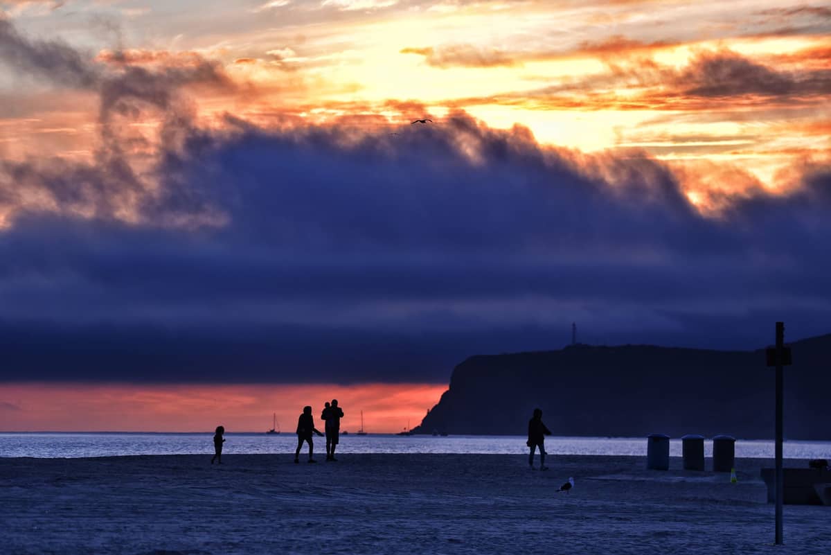 Menschen, die bei Sonnenuntergang an einem breiten Sandstrand spazieren, mit der markanten Silhouette von Point Loma im Hintergrund.