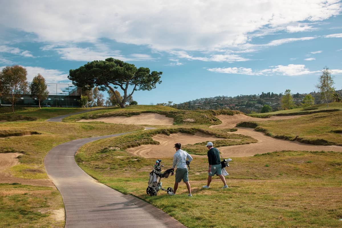 Zwei Golfspieler, die auf einem hügeligen Golfplatz einen gewundenen Pfad entlanggehen, unter einem blauen Himmel mit vereinzelten Wolken, mit entfernten Häusern und Bäumen im Hintergrund.