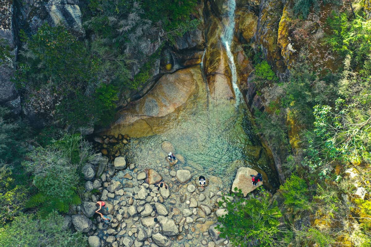 Luftaufnahme eines abgelegenen felsigen Wasserfalls und eines Pools, umgeben von dichtem Grün, mit einigen Wanderern, die das Gebiet darunter erkunden.