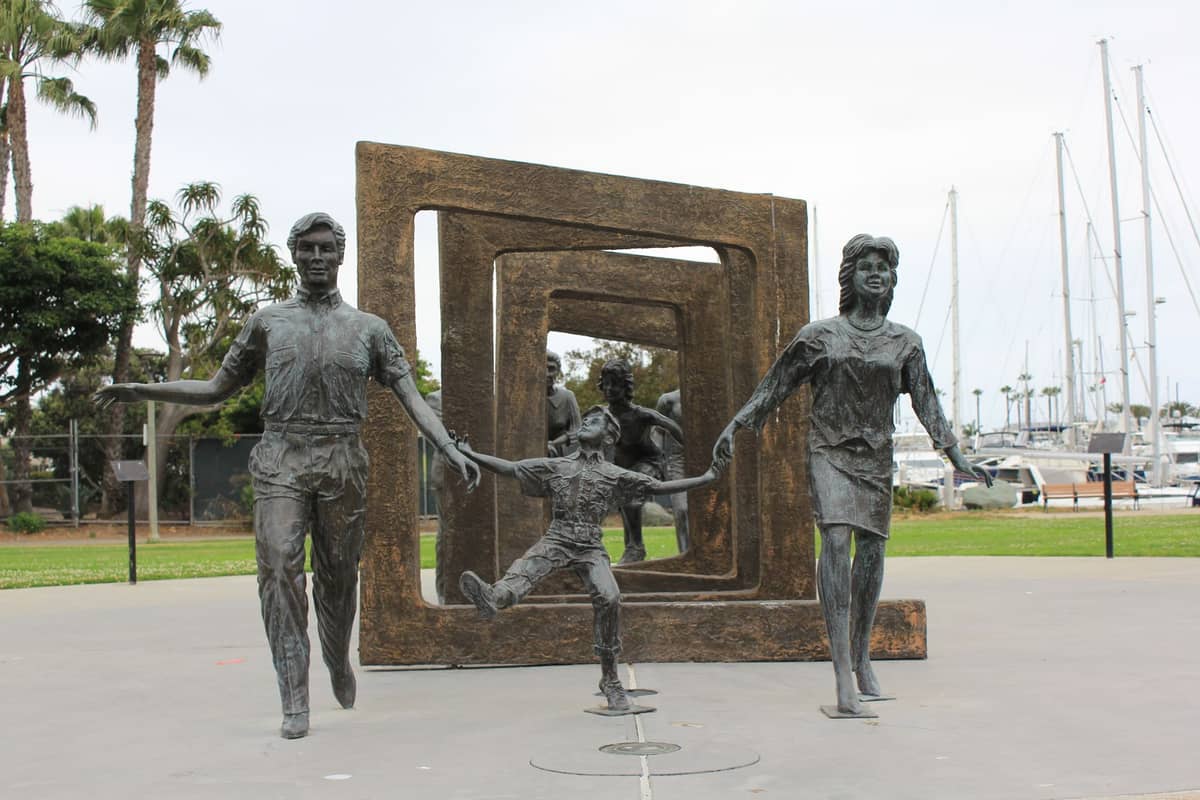 Bronzeskulpturen einer Familie in Bewegung im Freien, mit einem Yachthafen und Booten im Hintergrund und geometrischen Kunstrahmen dahinter.