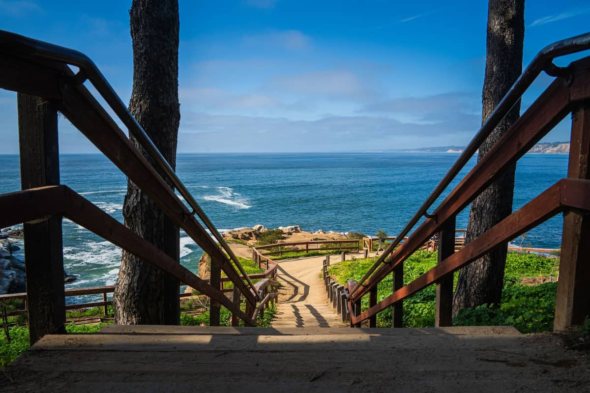 Blick die Holztreppe an der La Jolla Cove in San Diego hinunter, die zu Meereskliffs und blauem Wasser unter klarem Himmel führt.