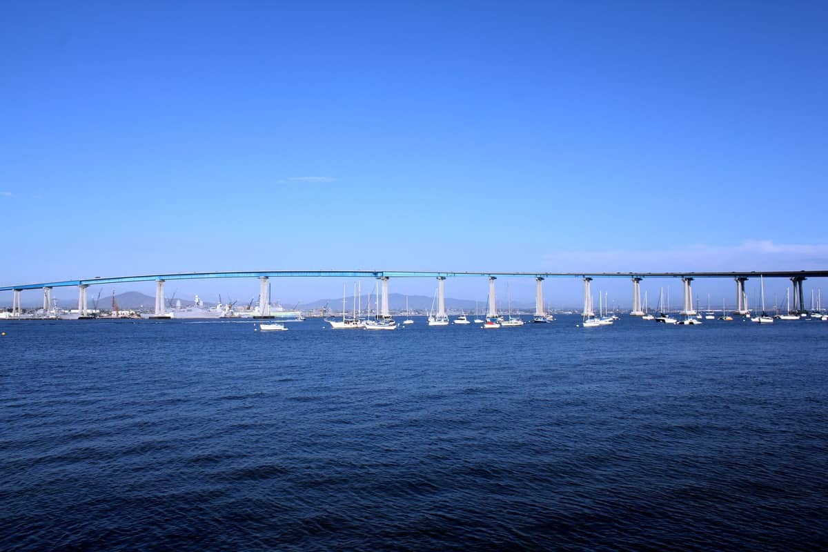 Breite horizontale Ansicht der Coronado Bridge, die sich über die San Diego Bay wölbt, mit Segelbooten auf dem Wasser unter einem klaren blauen Himmel.