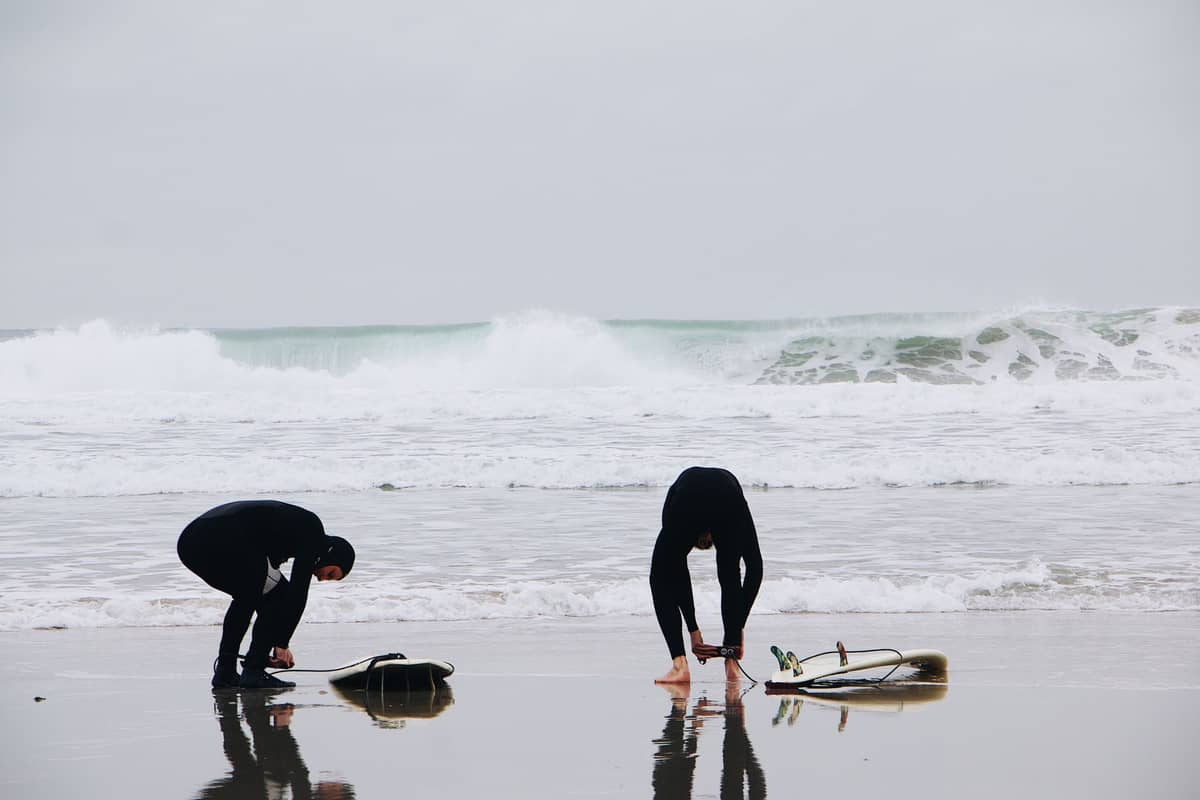 Zwei Surfer in Neoprenanzügen stehen an einem bewölkten Strand mit Surfbrettern auf dem nassen Sand und rauen Wellen im Hintergrund.