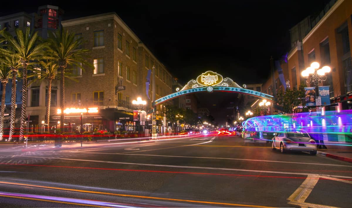 Nachtaufnahme des Eingangsbogens von San Diegos Gaslamp Quarter, belebte Straße mit Palmen, Neonlichtern und verwischten Lichtspuren von Autos.