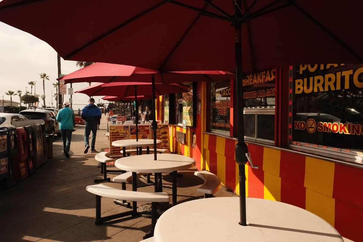 พื้นที่นั่งรับประทานอาหารกลางแจ้งของร้านทาโก้บรรยากาศสบายๆ ใน San Diego พร้อมร่มสีแดง กำแพงสีเหลืองสดใสและสีแดง และผู้คนที่เดินผ่านไปมา