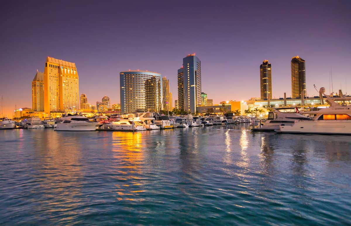Die Skyline von San Diego bei Sonnenuntergang mit Yachten im Hafen und dem Wasser im Vordergrund, Gebäude leuchten im Stadtlicht.