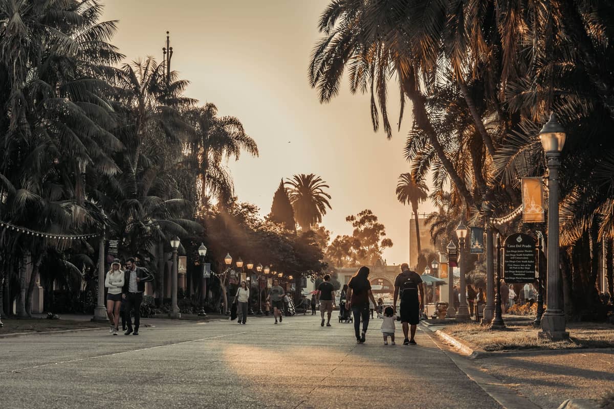 Menschen spazieren bei Sonnenuntergang entlang der palmengesäumten Fußgängerpromenade im Balboa Park, San Diego, mit historischen Straßenlaternen und üppigem Grün.