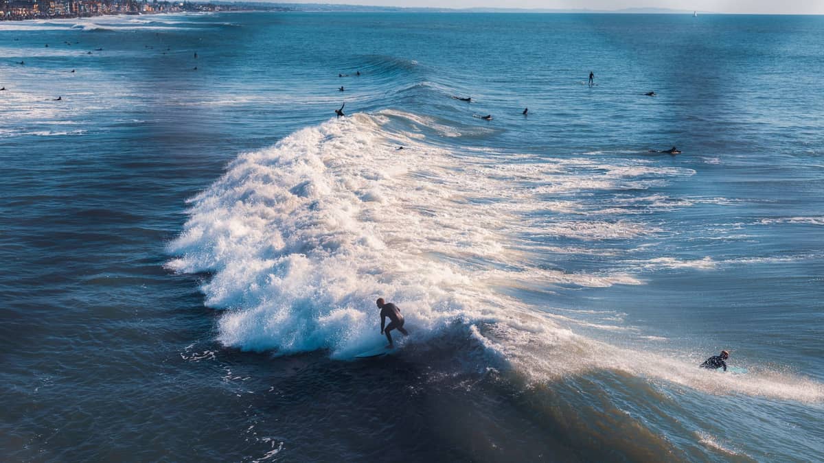 Surfer reiten und paddeln auf Wellen nahe einer belebten Küste, mit weißem Wasser und sichtbaren Surfbreaks unter klarem blauem Himmel.