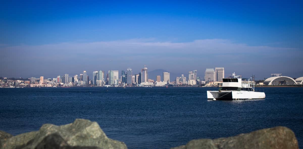 Weiter Blick auf die Skyline von San Diego, von der Bucht aus gesehen, mit einem weißen Boot auf dem Wasser und Felsen im Vordergrund.