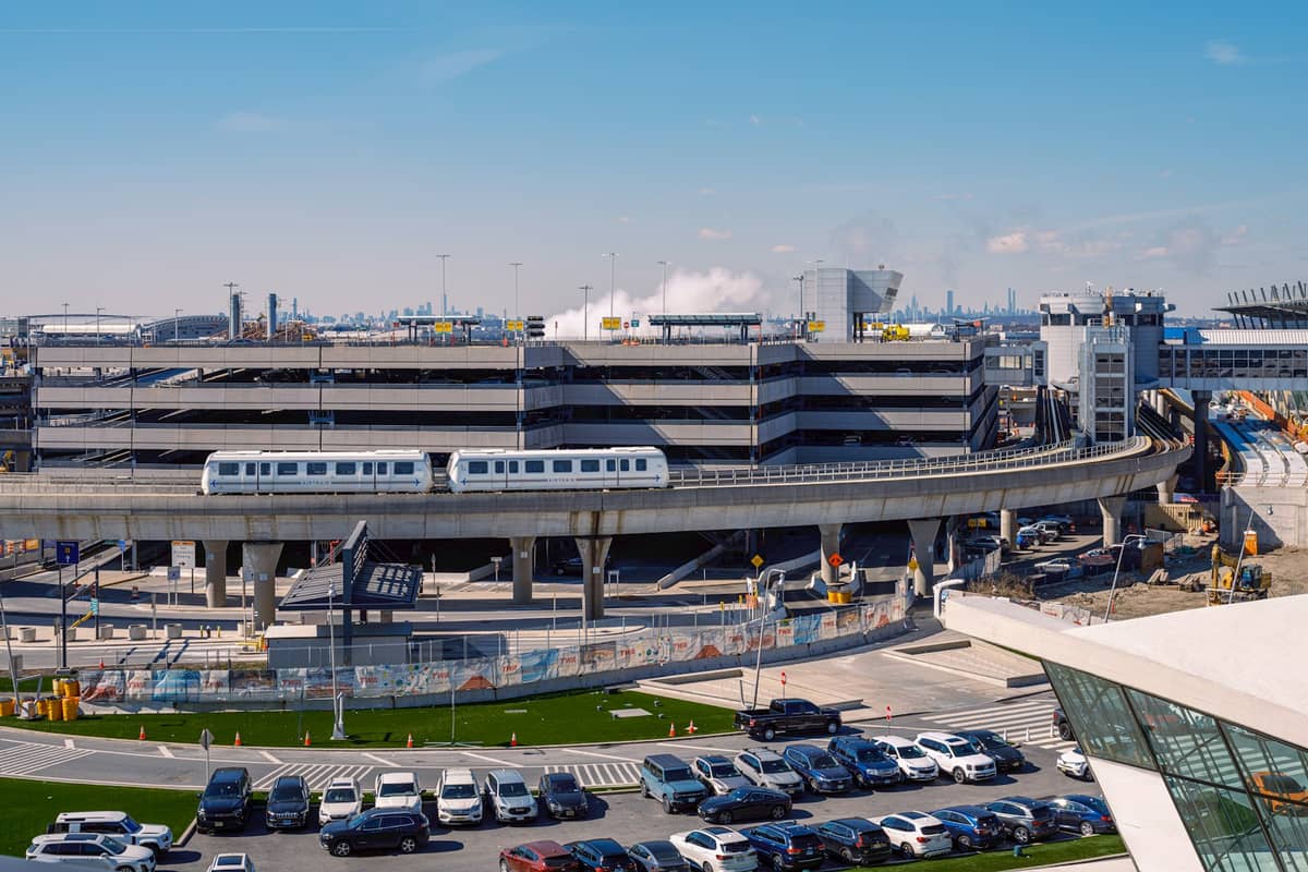 Großes mehrstöckiges Flughafenparkhaus mit Autos, AirTrain und Terminalgebäuden unter einem klaren blauen Himmel an einem großen Flughafen.