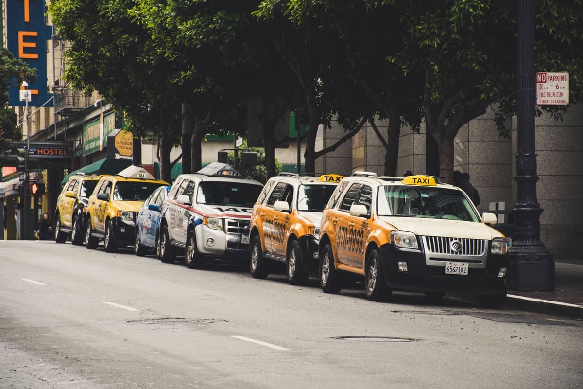 Eine Reihe von San Francisco-Taxis, die entlang einer Stadtstraße aufgereiht sind und in der Nähe von Stadtgebäuden und Grünflächen auf Fahrgäste warten.