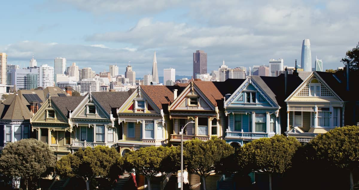 Klassische Reihe bunter Victorian Painted Ladies in San Francisco mit der Skyline der Stadt im Hintergrund, Bäume und Straße im Vordergrund.
