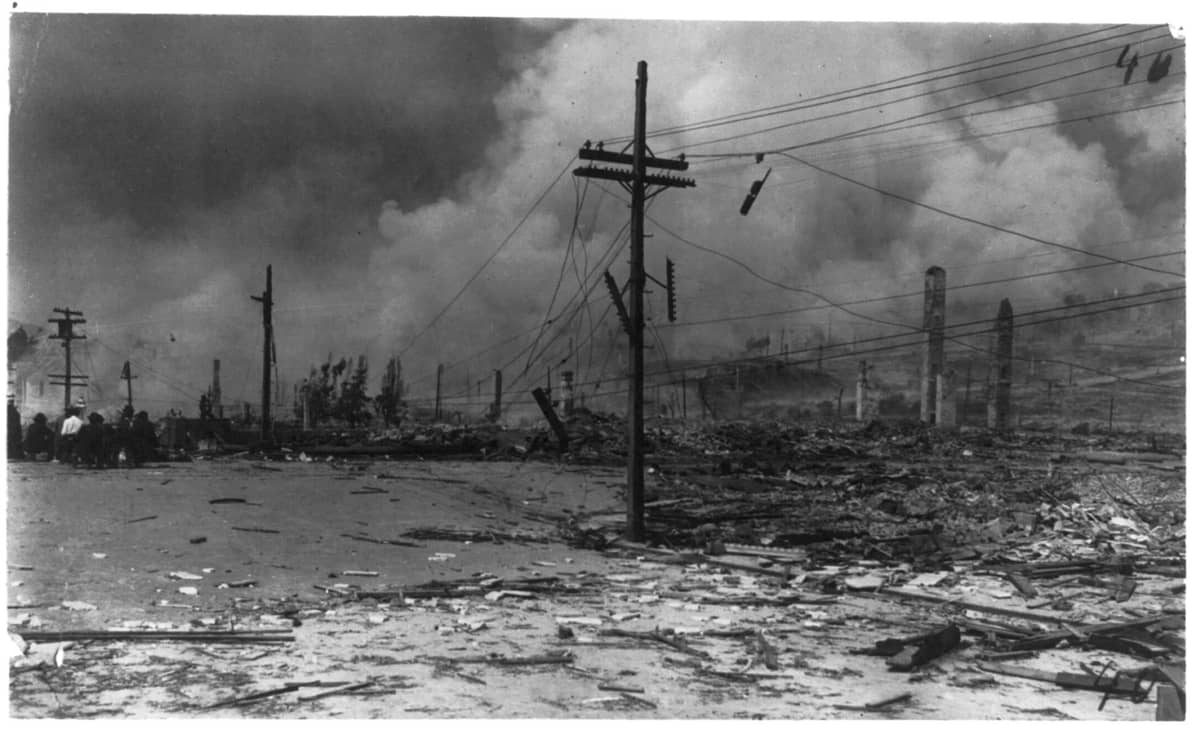 Historisches Schwarz-Weiß-Foto, das weitreichende Zerstörung und Rauch nach dem Erdbeben von San Francisco 1906 zeigt, mit verstreuten Trümmern und beschädigten Versorgungsmasten.