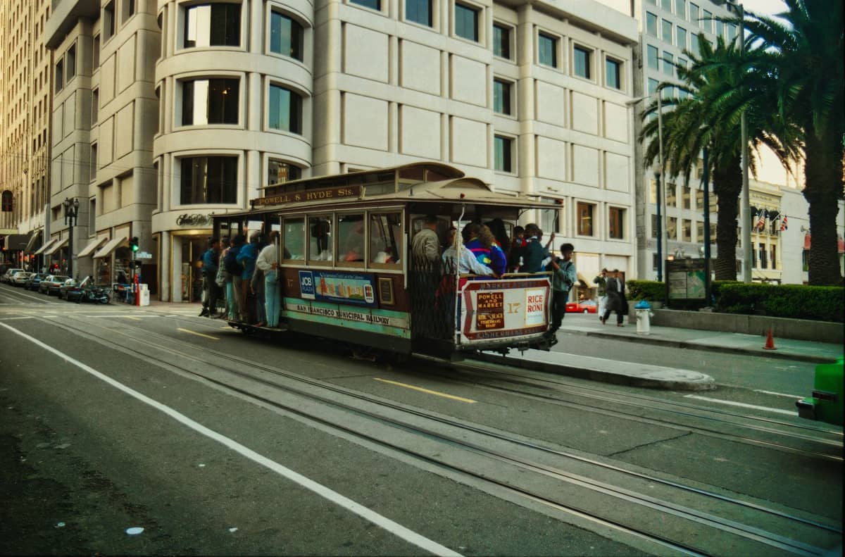 Eine klassische Cable Car aus San Francisco voller Fahrgäste fährt eine Steigung hinauf, im Hintergrund Palmen und Stadtgebäude.