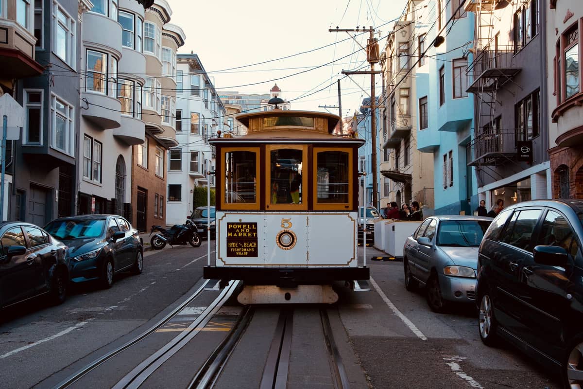Eine Seilbahn in San Francisco auf Schienen, umgeben von Autos und bunten Gebäuden auf einer Stadtstraße.