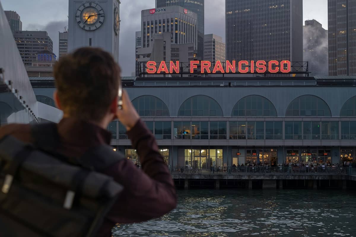 Osoba fotografująca San Francisco Ferry Building z charakterystyczną wieżą zegarową i neonowym szyldem widzianym od strony nabrzeża.