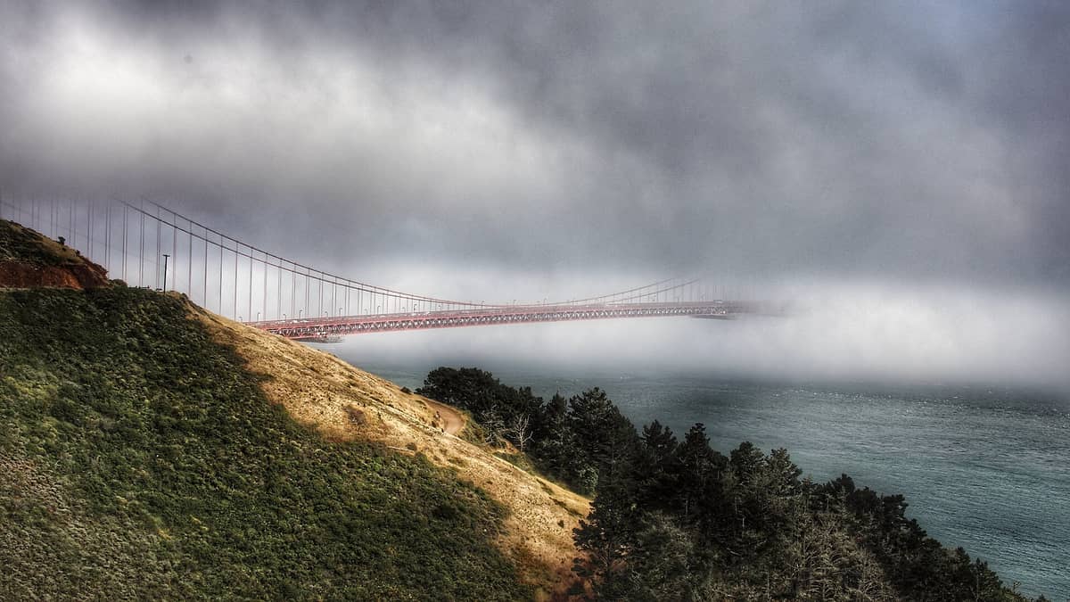 Die Golden Gate Bridge, die sich in dichten Meeresnebel erstreckt, von einem Hügel aus gesehen mit dramatisch bewölktem Himmel und geringer Sicht über dem Wasser.