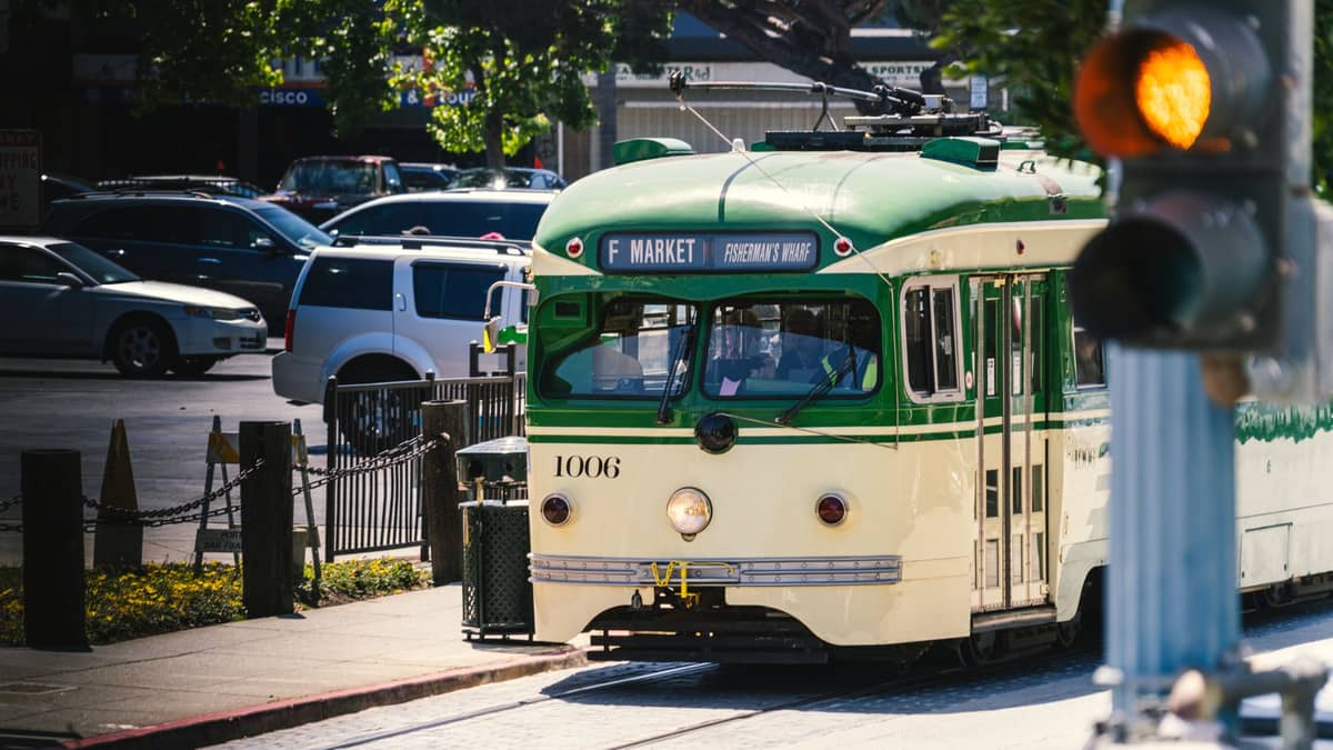 Histórico tranvía de la línea F Market en San Francisco se acerca a una parada con tráfico y árboles visibles al fondo.