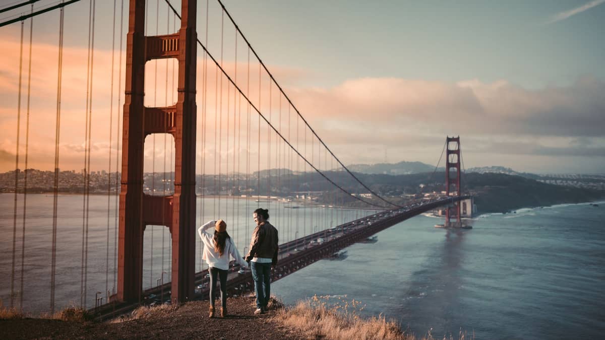 Una pareja tomada de la mano en un mirador sobre el puente Golden Gate al atardecer en San Francisco, con nubes dramáticas y la ciudad al fondo.