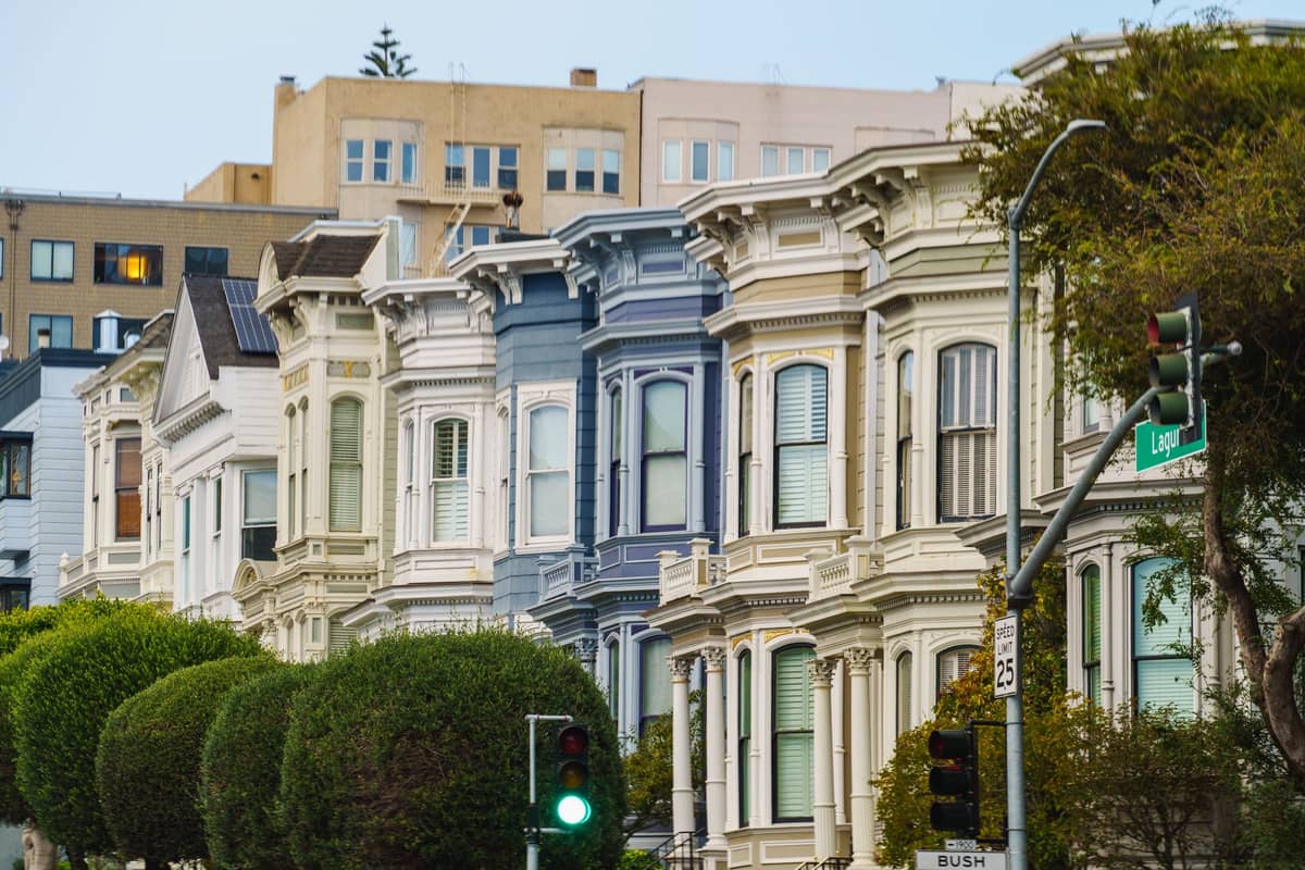 Reihe klassischer viktorianischer Häuser in Pastellfarben in einer hügeligen Straße in San Francisco mit belaubten Bäumen und Stadtgebäuden im Hintergrund.