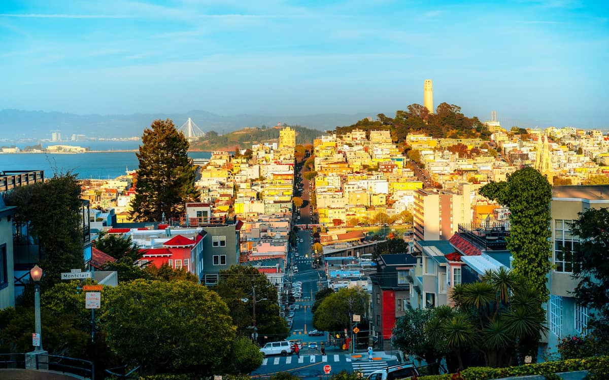 Sonnendurchflutete Aussicht eine hügelige Straße in San Francisco hinauf zur Coit Tower, mit der Bucht und der Brücke im Hintergrund, umgeben von klassischen Stadtgebäuden und Grünflächen.