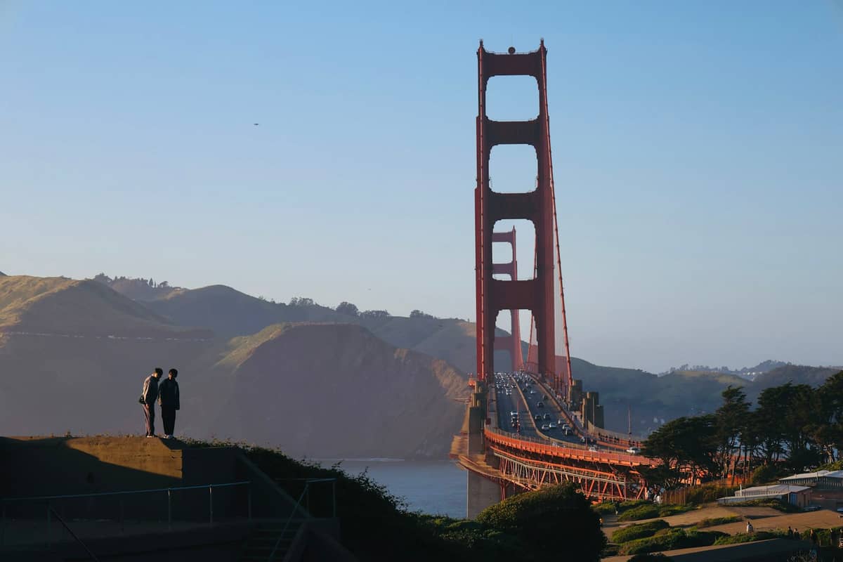 Zwei Personen stehen bei Sonnenaufgang oder Sonnenuntergang neben der Golden Gate Bridge mit der Bucht und sanften Hügeln im Hintergrund.