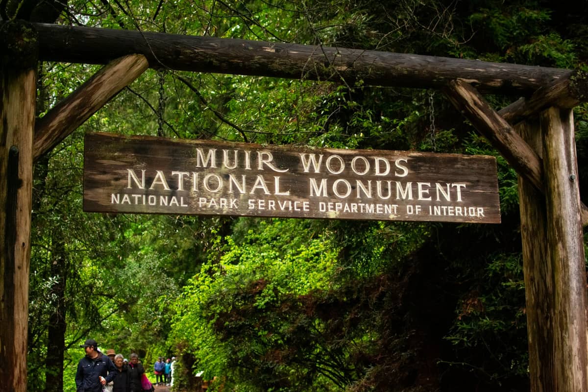 Hölzernes Eingangsschild des Muir Woods National Monument, umgeben von hohen Redwoods und Grünpflanzen, mit Menschen, die unter dem Schild entlanggehen.