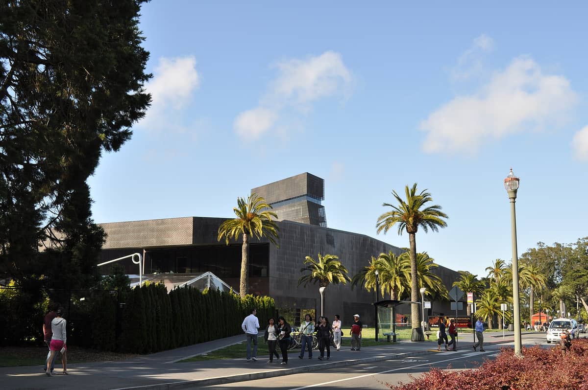 Vista exterior do de Young Museum no Golden Gate Park, com pessoas caminhando e palmeiras na frente.