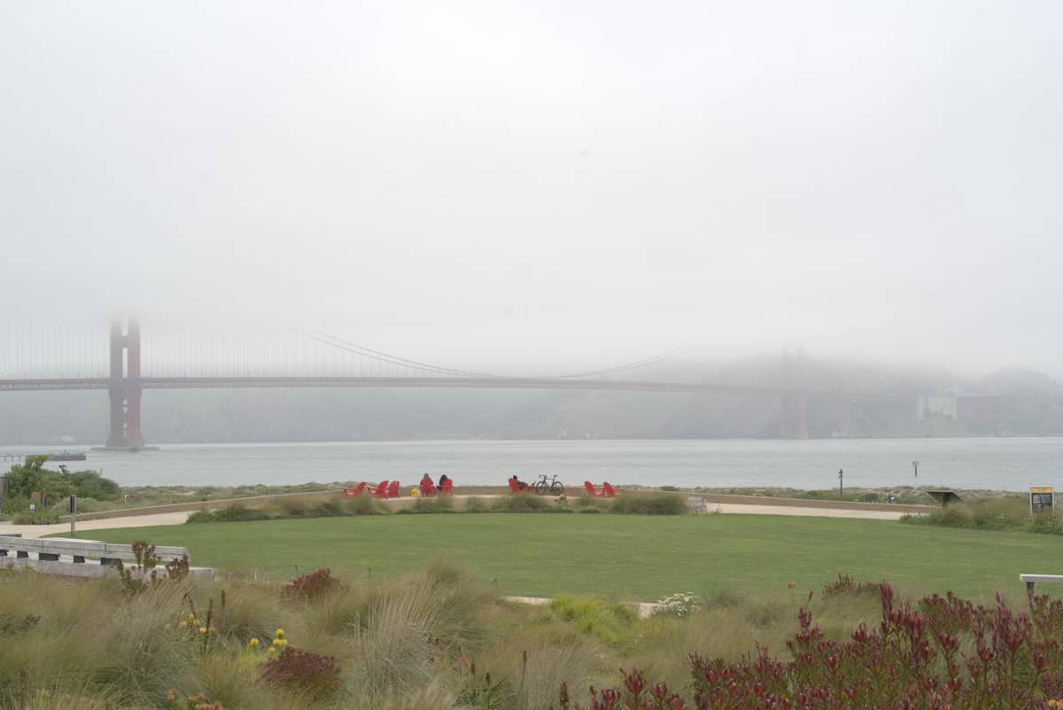 Neblina cobre a Golden Gate Bridge e os parques ao redor, com céu nublado e baixa visibilidade, refletindo as condições típicas de verão em San Francisco.