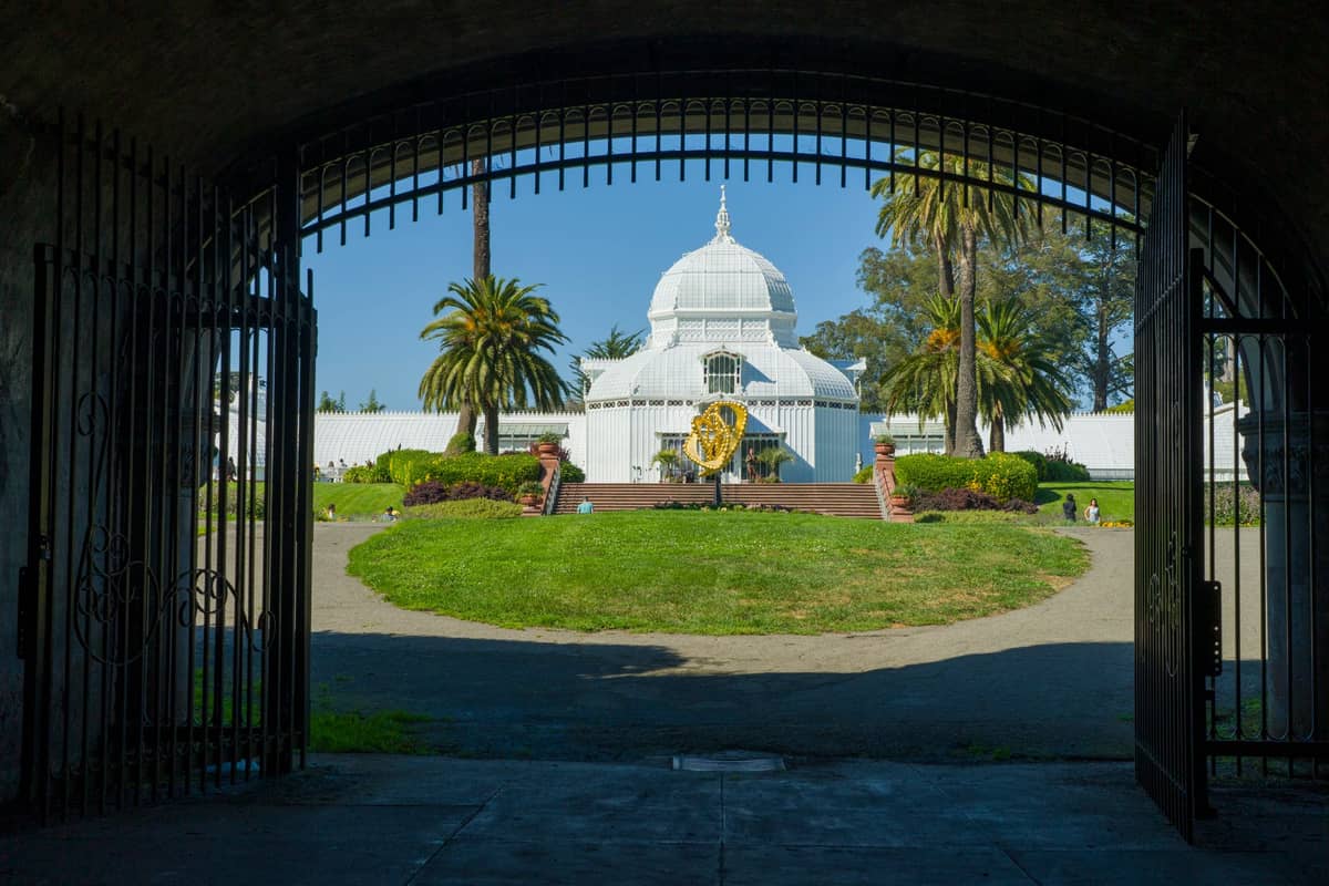 Vista através de um portão em arco em direção ao Conservatory of Flowers de vidro branco cercado por palmeiras e vegetação no Golden Gate Park.