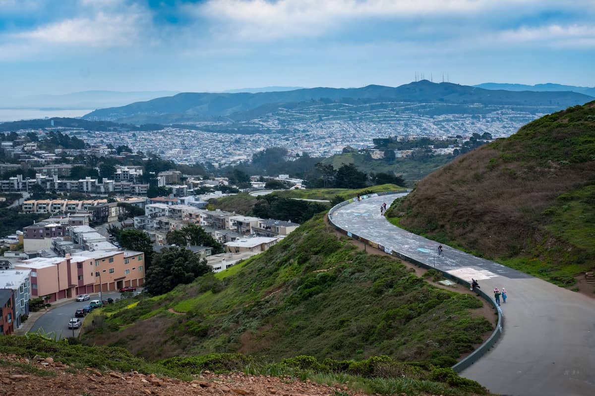 Blick von einem Hügelpark in San Francisco mit einem geschwungenen Weg, grünen Hängen und einem weiten Stadtpanorama unter bewölktem Himmel.