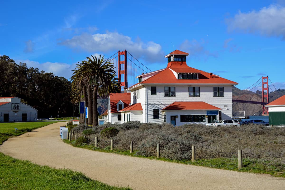 Piaszczysty szlak zakręca obok białego budynku z czerwonym dachem, palmy i widocznego w tle Golden Gate Bridge.