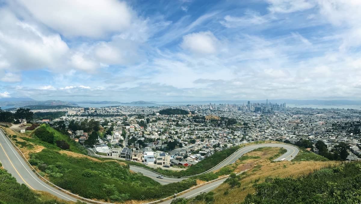 Szeroka panorama z wzgórza w San Francisco przedstawiająca krętą drogę, panoramę miasta i linię horyzontu pod błękitnym niebem z chmurami.