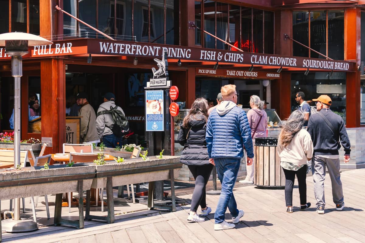 Personas caminando cerca de un animado restaurante frente al mar con letreros que anuncian fish and chips y clam chowder en San Francisco.