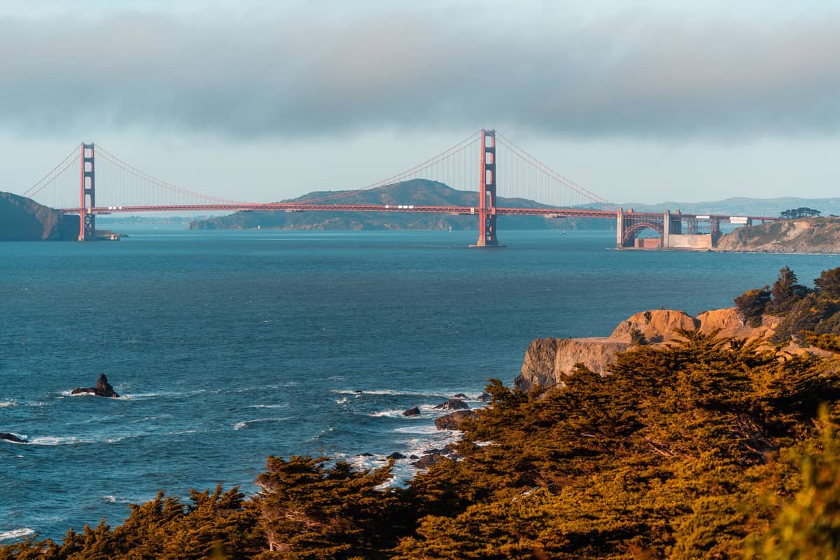 Weite Aussicht auf die Golden Gate Bridge mit der Pazifikküste, felsigen Klippen und Bäumen im Vordergrund in San Francisco.