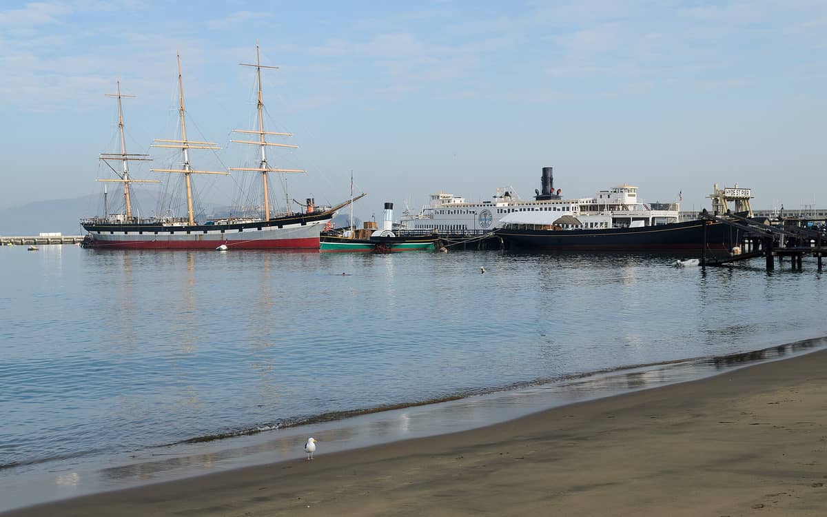 Barcos históricos, entre ellos el Balclutha y el Eureka, amarrados en el Hyde Street Pier del Parque Histórico Nacional Marítimo de San Francisco, con aguas tranquilas y una orilla arenosa en primer plano.