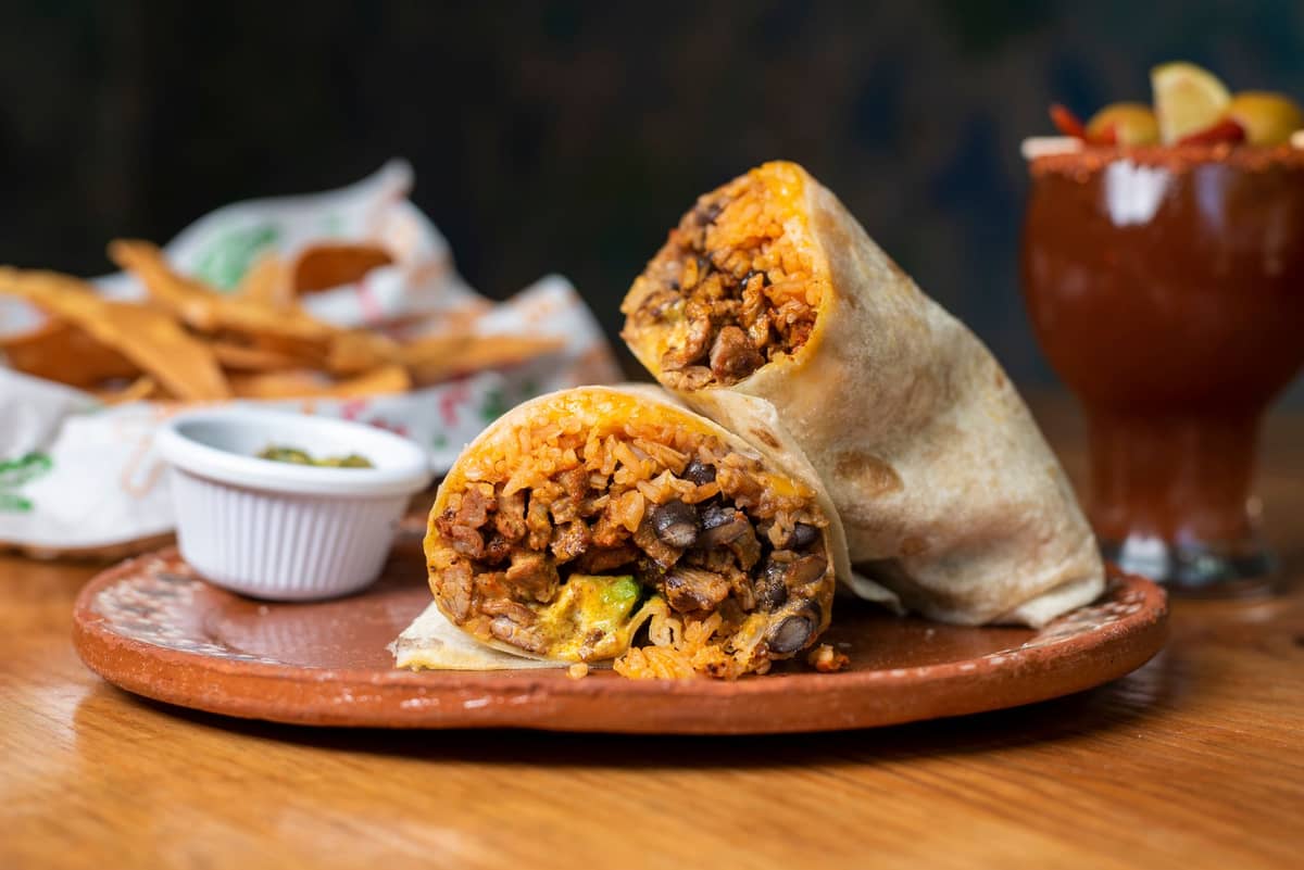 Duże burrito przekrojone na pół ukazujące ryż, fasolę i mięso w środku, na glinianym talerzu z nachosami i salsą w tle.