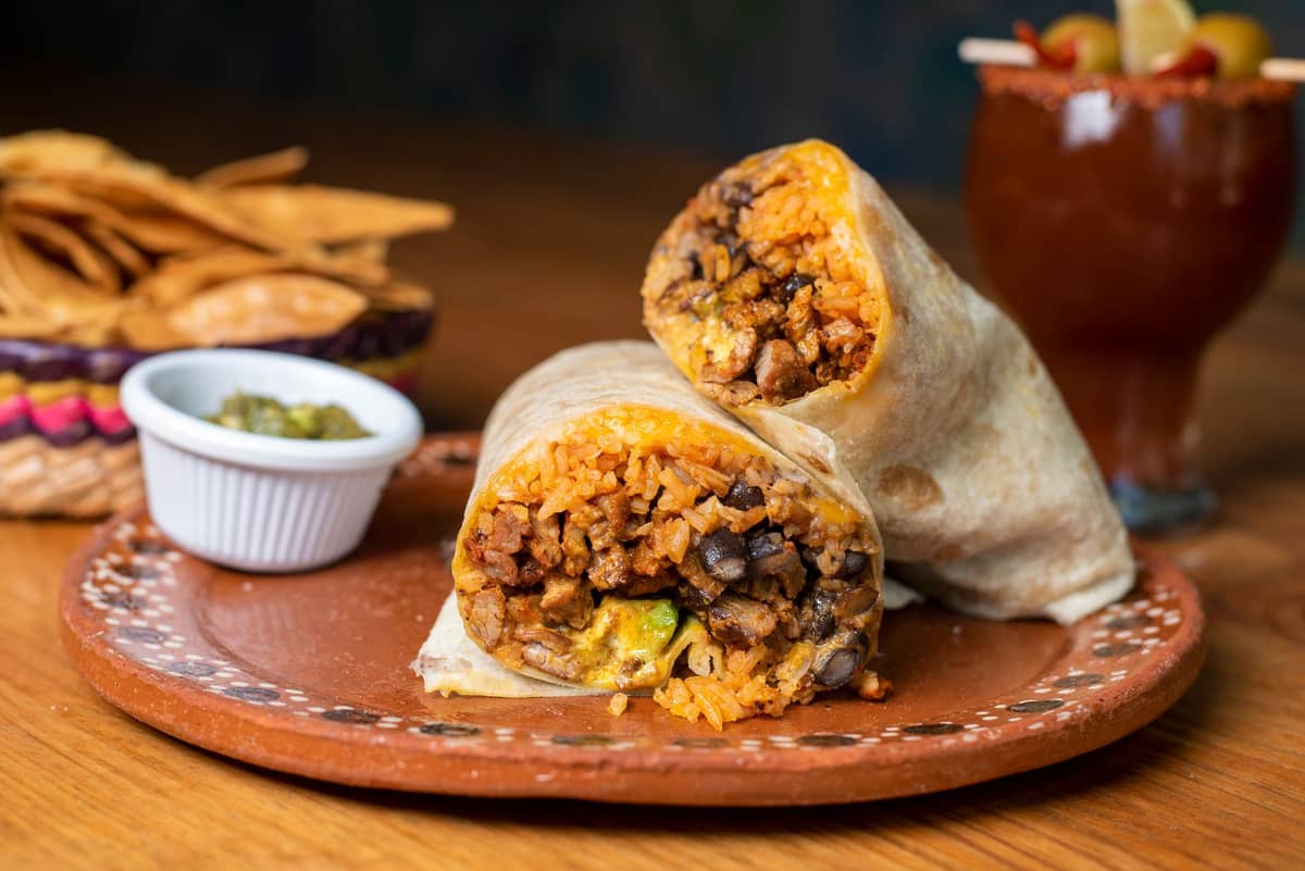 Un burrito estilo Mission cortado por la mitad en un plato decorativo, con arroz, frijoles y carne visibles, acompañado de totopos, salsa y una bebida.