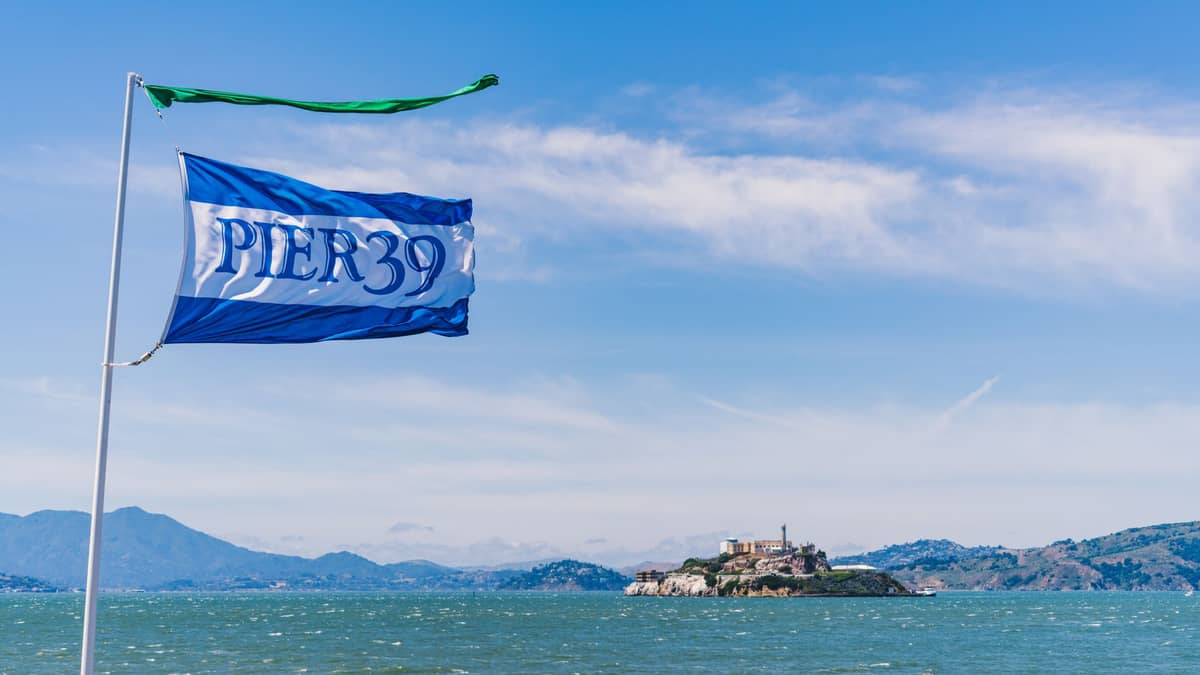Eine Pier 39-Flagge weht im Vordergrund, mit Alcatraz Island und der San Francisco Bay im Hintergrund unter einem klaren blauen Himmel.