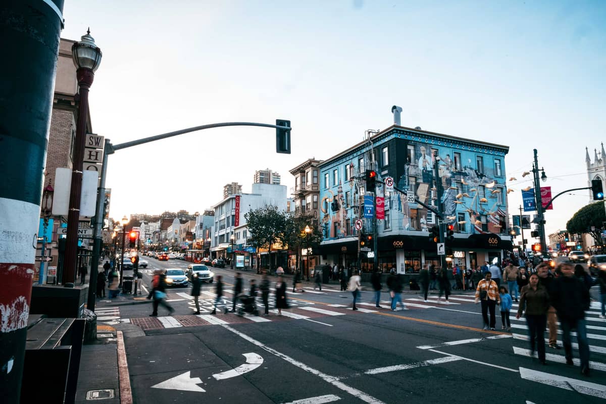 Kerumunan orang menyeberangi jalan sibuk di kawasan North Beach, San Francisco, dengan mural berwarna-warni dan tempat hiburan malam yang mudah dikenali saat senja.