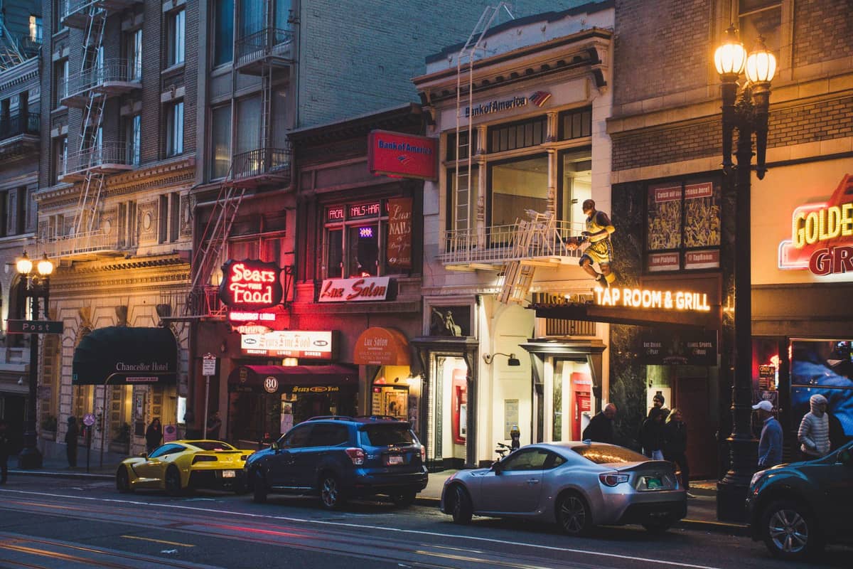 Una animada calle de San Francisco al atardecer con letreros de neón encendidos, aceras llenas de gente y personas entrando a bares y clubes nocturnos.