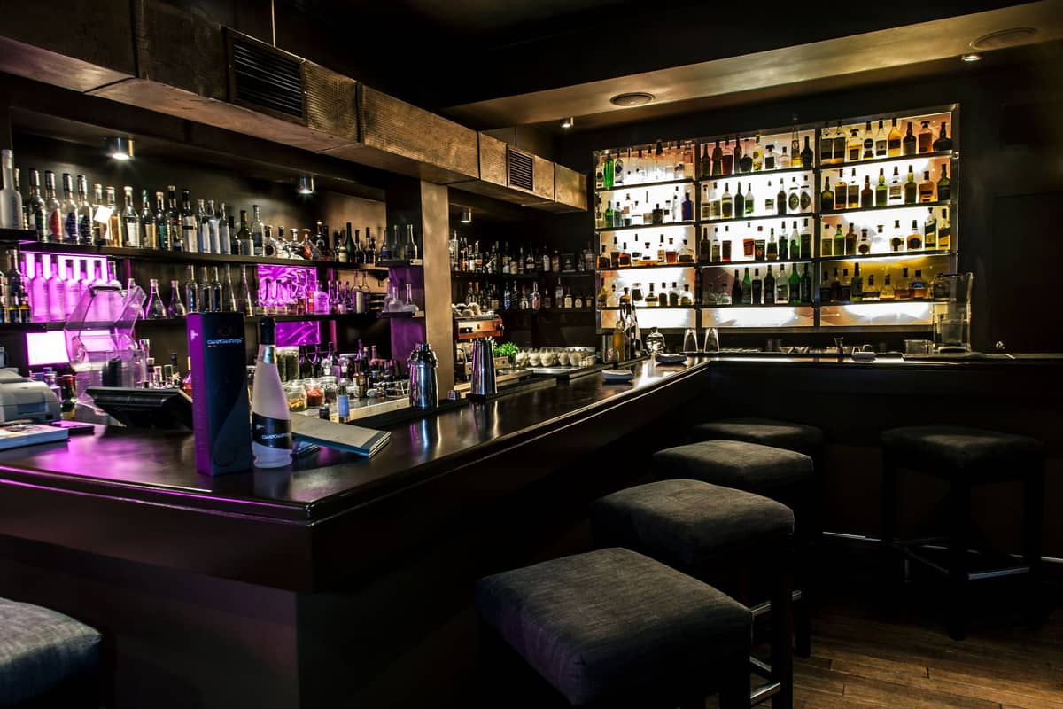 Interior bar koktail modern dengan pencahayaan redup, rak-rak berisi botol, dan beberapa bangku bar di sekitar meja bar sudut.