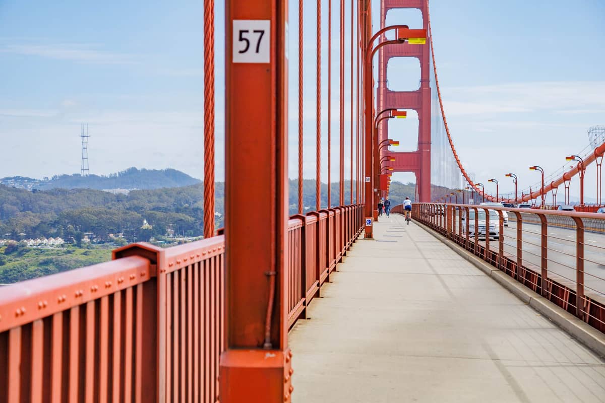 Jalur pejalan kaki Golden Gate Bridge dengan pemandangan jelas menara-menara ikoniknya dan beberapa orang berjalan di hari yang cerah.