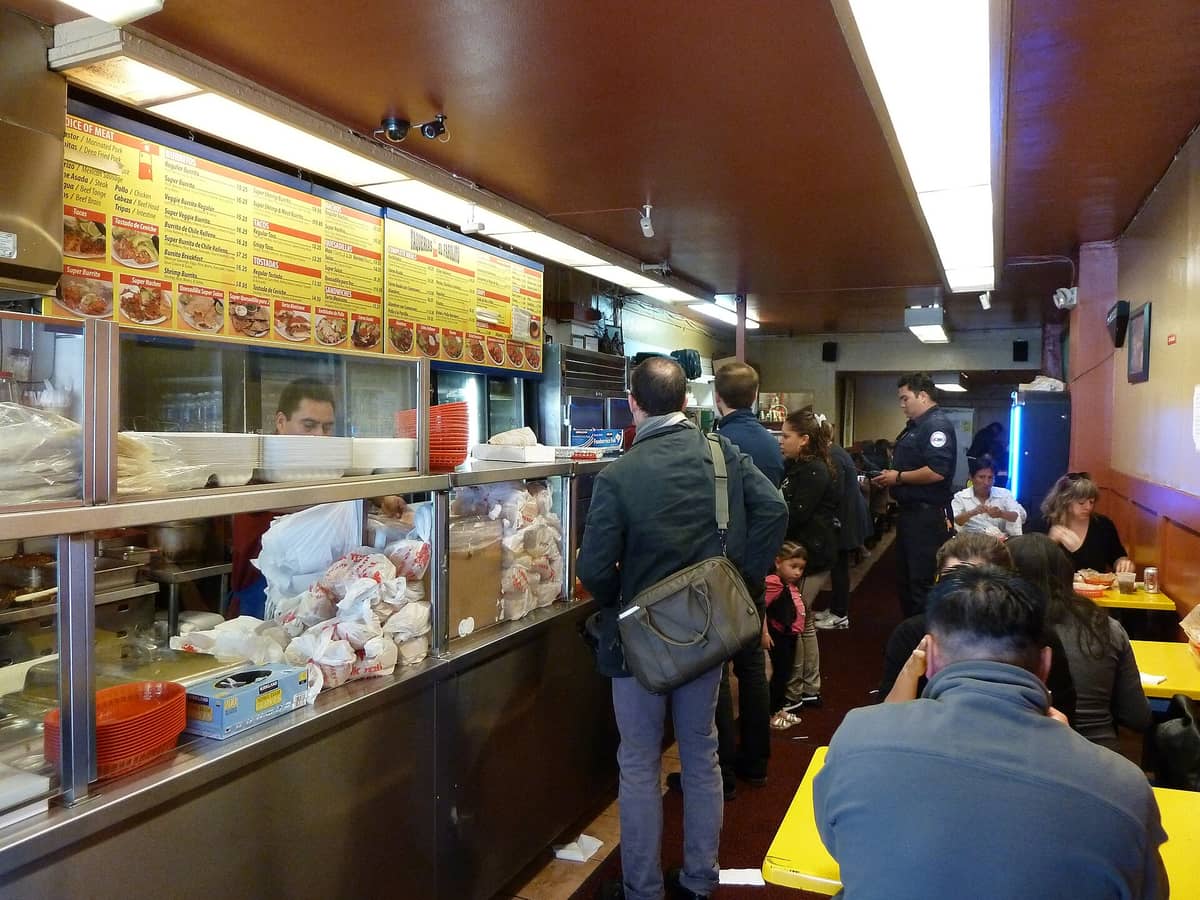 Interior sebuah taqueria ramai di San Francisco dengan orang-orang yang mengantri untuk memesan dan makan di meja-meja berwarna kuning.
